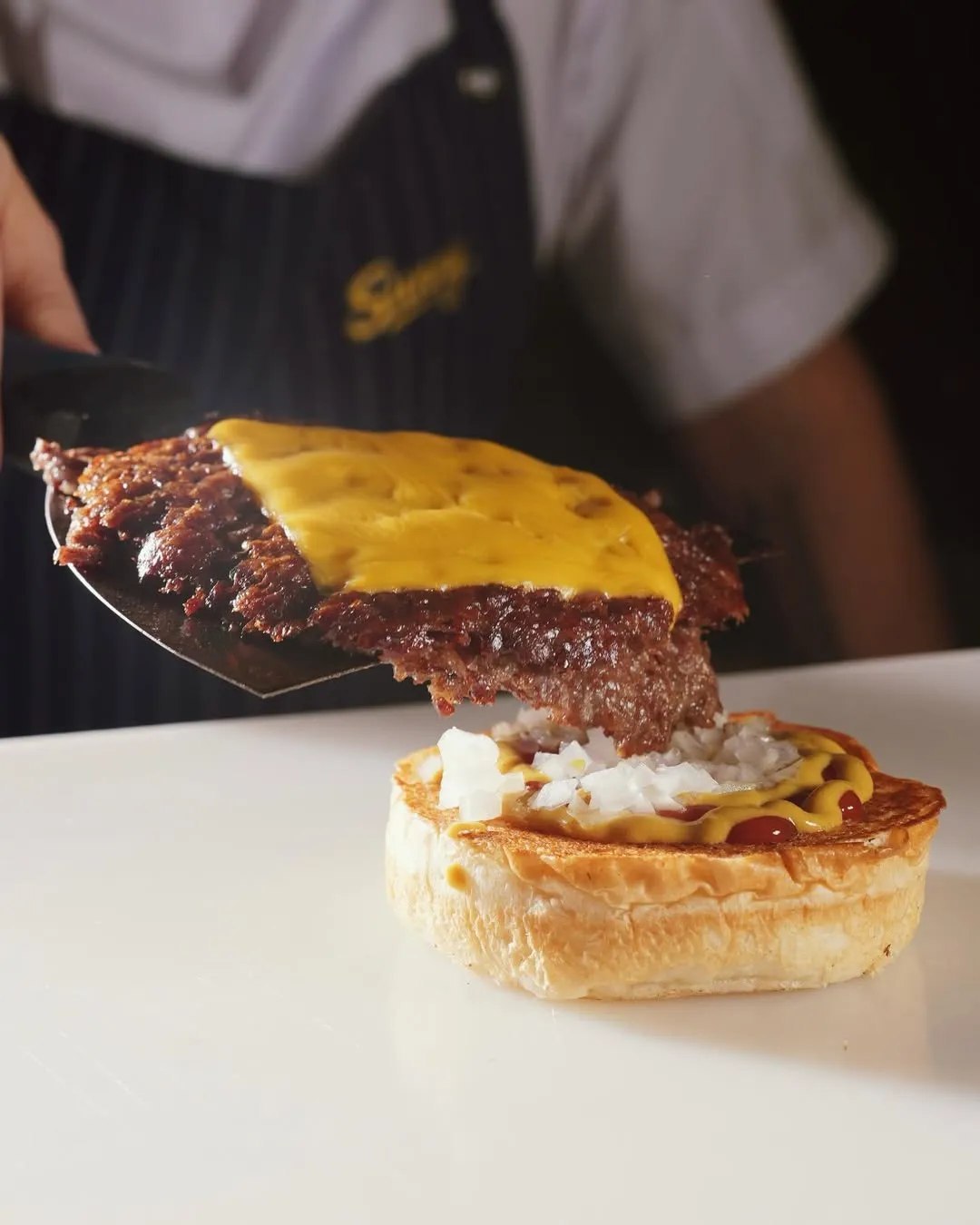 Beyaz zeminde parlak ekmekli, iki köfteli, erimiş peynirli, domatesli ve marullu smash burger; yanında kâğıttan taşan çıtır patates, sarı sos ve Sunny markalı limonata.