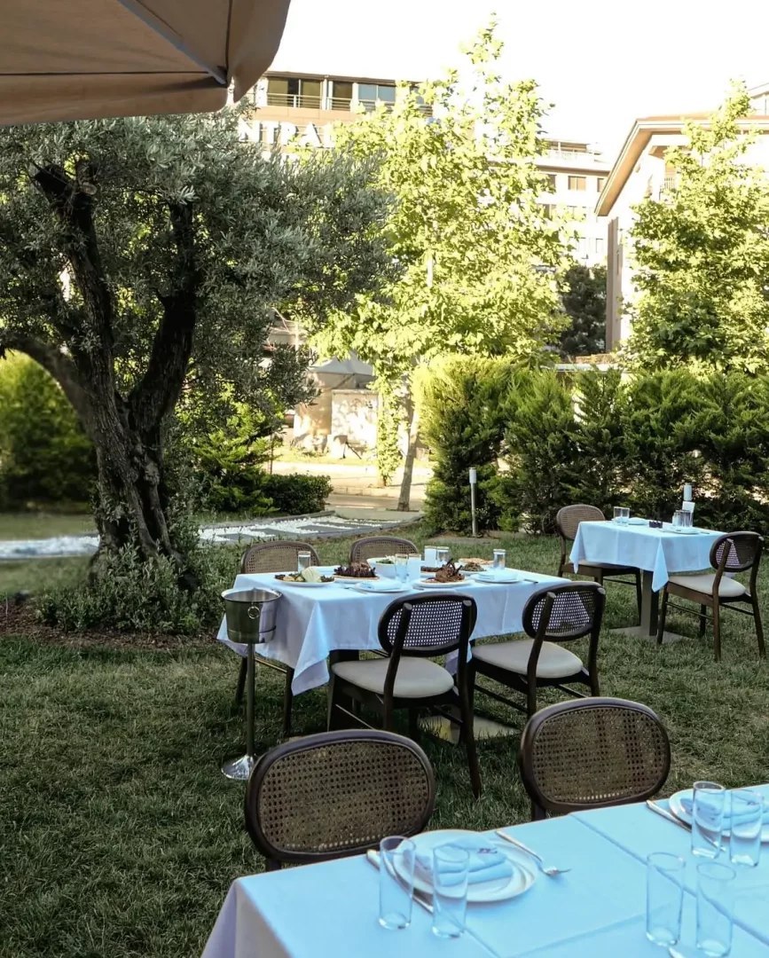 Etiler'deki Tekkebapçı restoranının zeytin ağaçları ve yeşil bitki örtüsüyle çevrili bahçesinde, beyaz masa örtüleri serilmiş ve üzerinde tabaklar ile bardaklar bulunan, bazı masalarda yiyeceklerin de yer aldığı, misafirlerini bekleyen şık ve ferah bir açık hava yemek alanı.